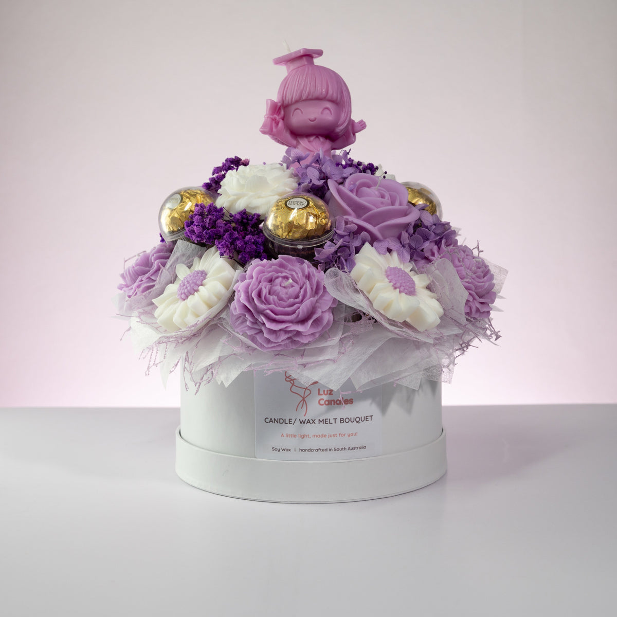 Luz Candles Au - A decorative candle and wax melt bouquet arranged in a round white gift box. The textured purple petals, gold foil-wrapped candies, and a small purple prom doll figure on top.