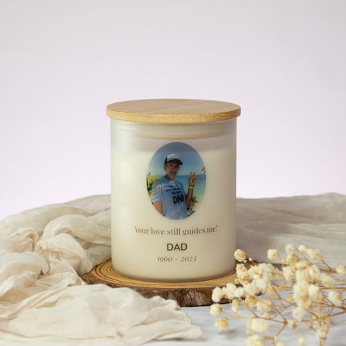 Luz Candles Au - A personalized jar candle with a bamboo lid sits on a wooden slice coaster, surrounded by draped cream fabric and dried baby's breath flowers. The candle features a custom photo of a man on the label with the text 'Your love still guides me! DAD 1960 - 2023'.