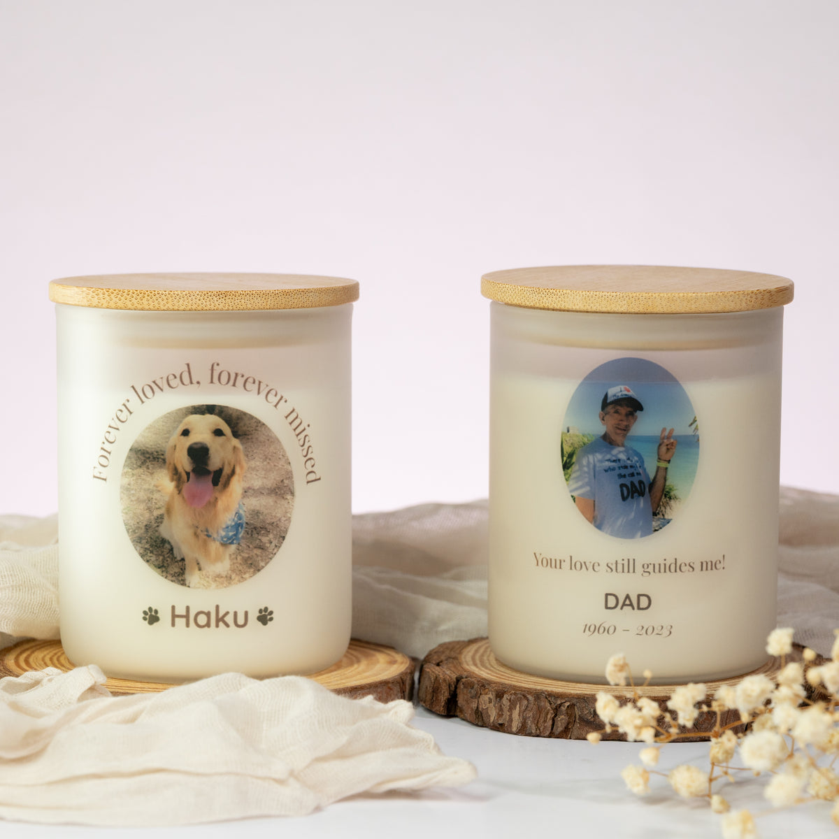 Luz Candles Au - Two personalized pet memorial candles in clear glass jars with bamboo lids, displayed on wood slices with soft fabric and dried flowers. One candle is labeled 'Forever loved, forever missed Haku' with a golden dog photo, and the other is labeled 'Your love still guides me DAD' with a man's photo and years '1960 - 2023'.