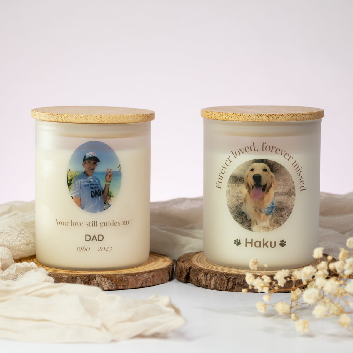 Luz Candles Au - Two personalized memorial candles in frosted glass jars with bamboo lids, displayed on wooden coasters with draped fabric and dried flowers. One candle is labeled 'DAD' with a photo of a man, and the other is labeled 'Haku' with a photo of a golden retriever.