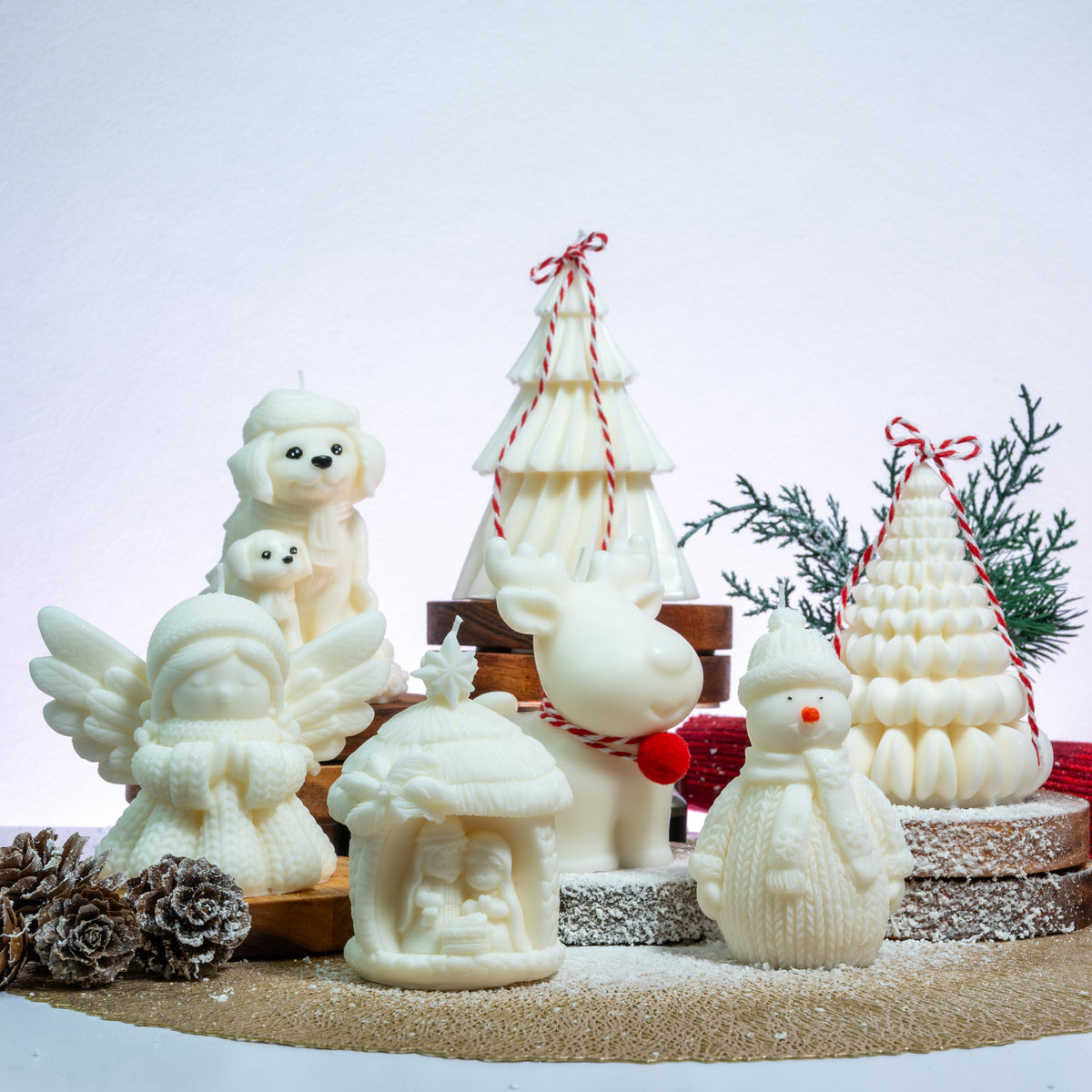 A set of white holiday-themed wax candles shaped like a dog and cat, an angel, a reindeer, a snowman, a house, and two Christmas trees, arranged on a rustic display with pinecones and greenery against a light background.