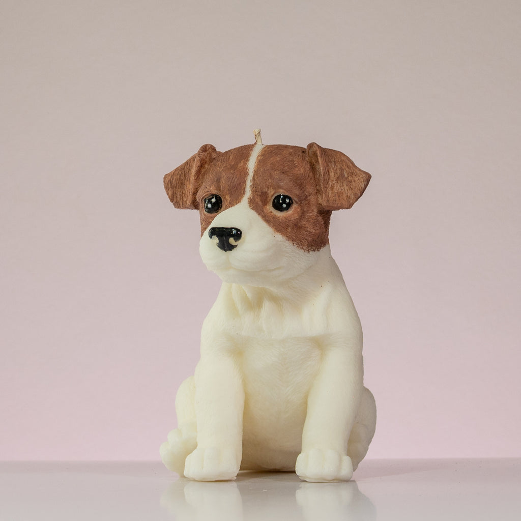 Luz Candles Au - A realistic dog-shaped candle, likely shaped like a Jack Russell Terrier, featuring brown and white coloring with black eyes and nose, sitting on a reflective white surface against a soft pink background.
