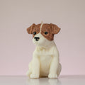 Luz Candles Au - A realistic dog-shaped candle, likely shaped like a Jack Russell Terrier, featuring brown and white coloring with black eyes and nose, sitting on a reflective white surface against a soft pink background.
