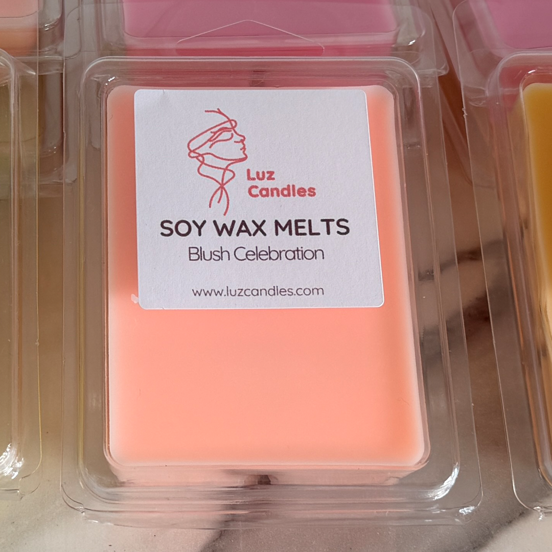 A square package of blush pink soy wax melts in a clear plastic clamshell container, with a white label reading 'Luz Candles' and 'SOY WAX MELTS Blush Celebration'.