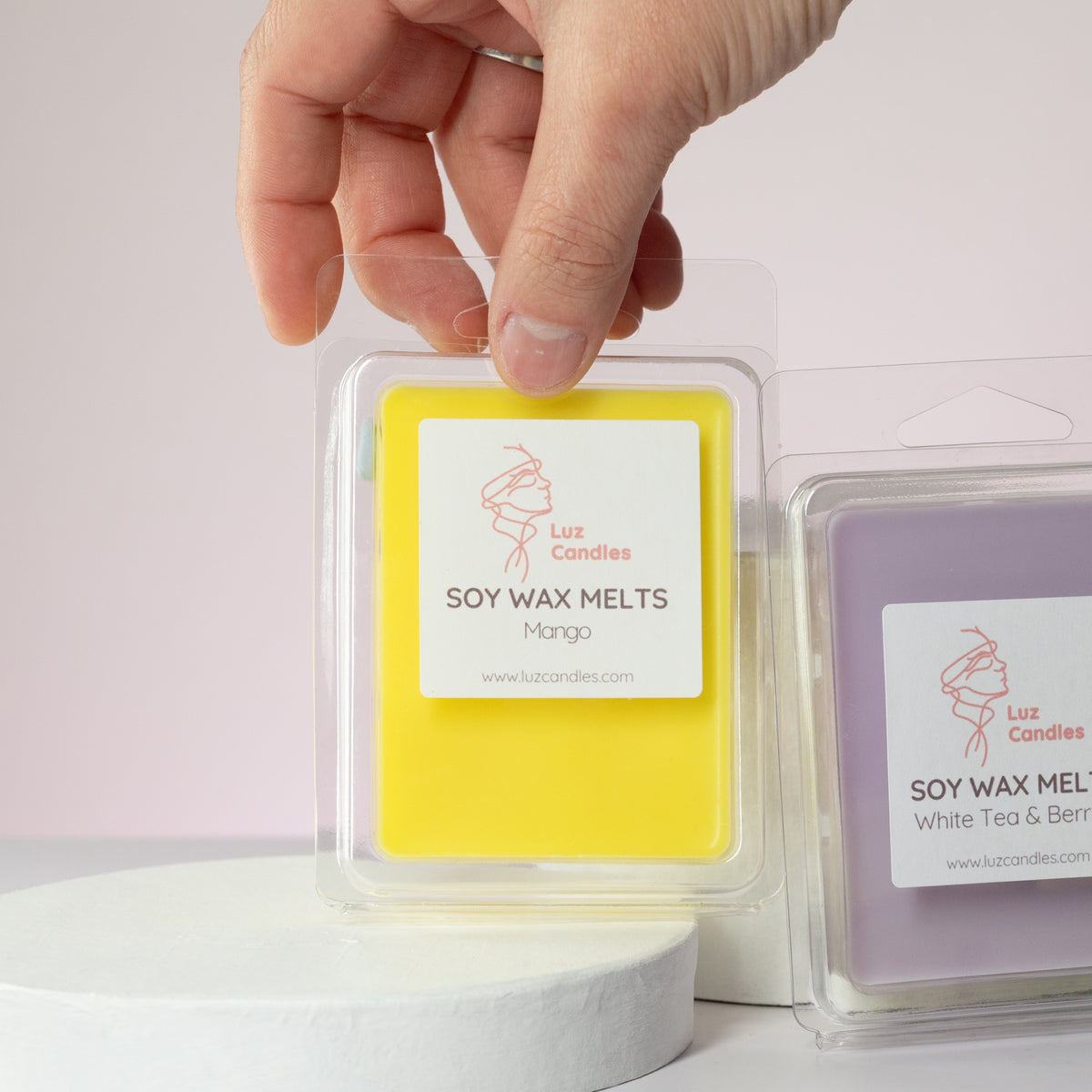 Luz Candles Au - A hand holds a clear plastic package of yellow soy wax melts labeled 'Luz Candles Soy Wax Melts Mango' in front of a lavender package on a white pedestal against a pale pink background.