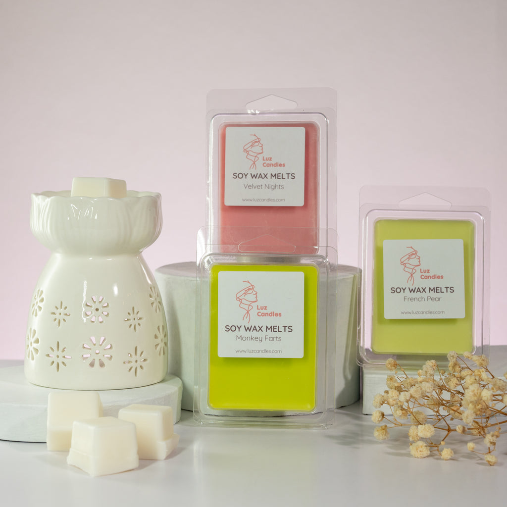 Luz Candles Au - A set of soy wax melts and a white ceramic diffuser with flower cutouts, arranged on a white surface against a pale pink background. The products include three packages of soy wax melts in pink, yellow-green, and white, along with several white wax cubes and dried floral decor.
