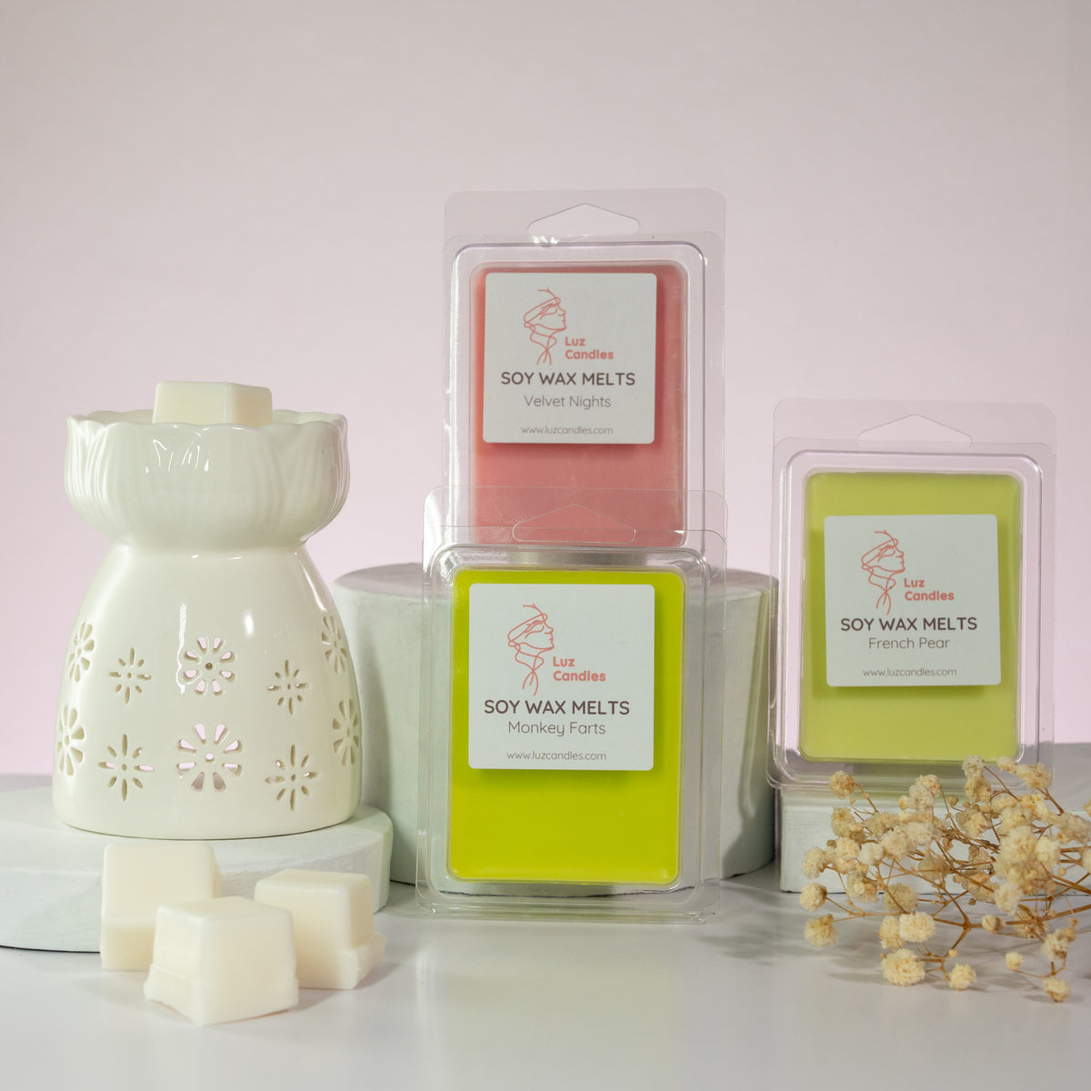Luz Candles Au - A set of soy wax melts and a white ceramic diffuser with flower cutouts, arranged on a white surface against a pale pink background. The products include three packages of soy wax melts in pink, yellow-green, and white, along with several white wax cubes and dried floral decor.