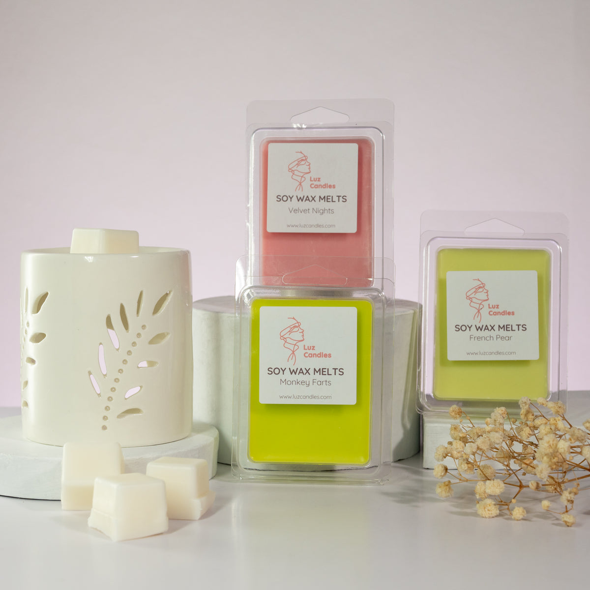 Luz Candles Au - Three packaged soy wax melts and one unboxed white wax melt with a decorative ceramic holder, arranged on a white surface against a pale pink background with dried flowers at the side.