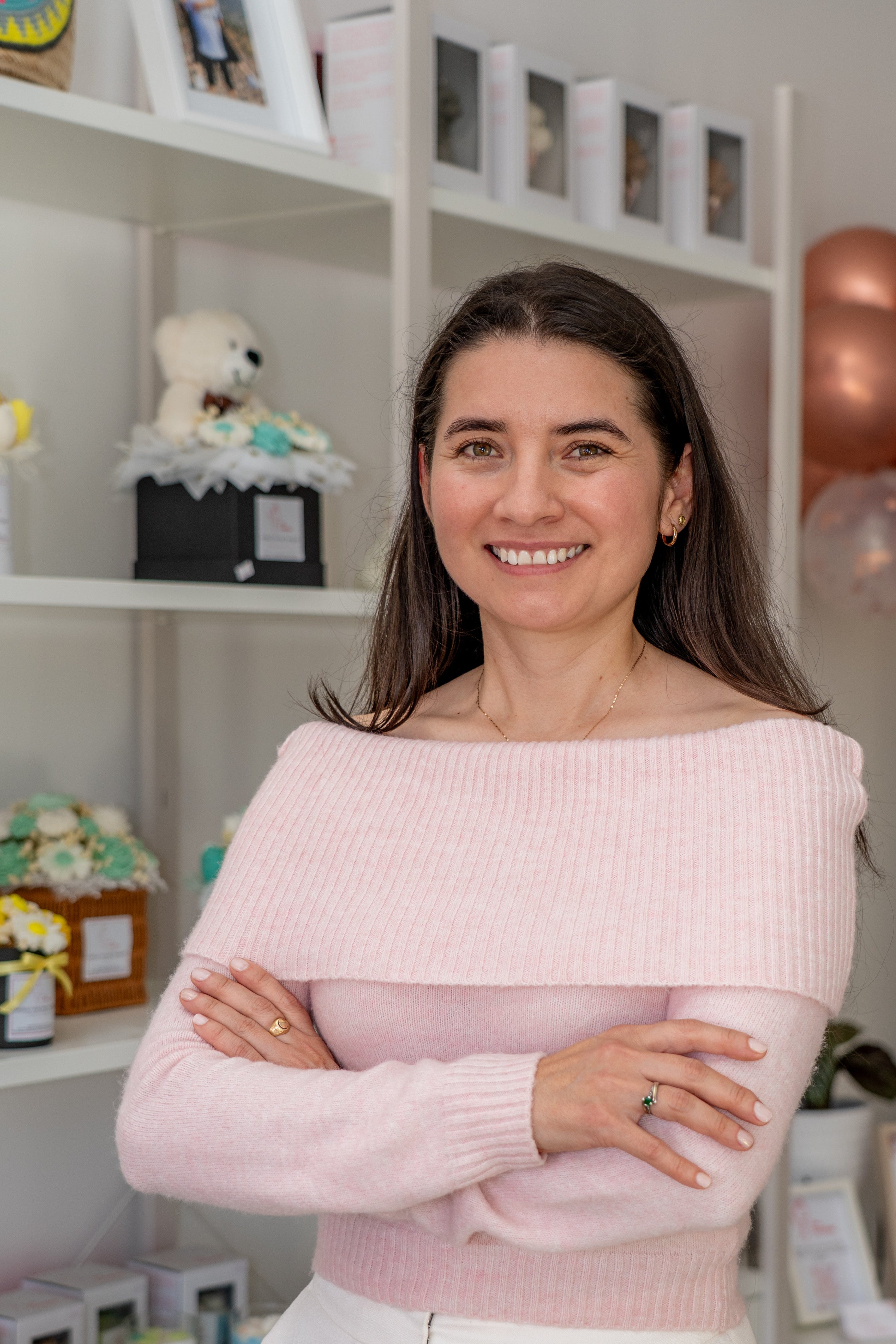 Luz smiling with dark hair stands indoors wearing a light pink off-the-shoulder knit sweater and white pants, with her arms crossed. The background shows white shelves with decorative items, framed pictures, balloons, and a small table.