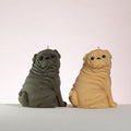 Luz Candles Au - Two pug-shaped candles sitting side by side on a white surface against a light pink background. The left candle is dark gray and the right candle is a warm beige/tan.