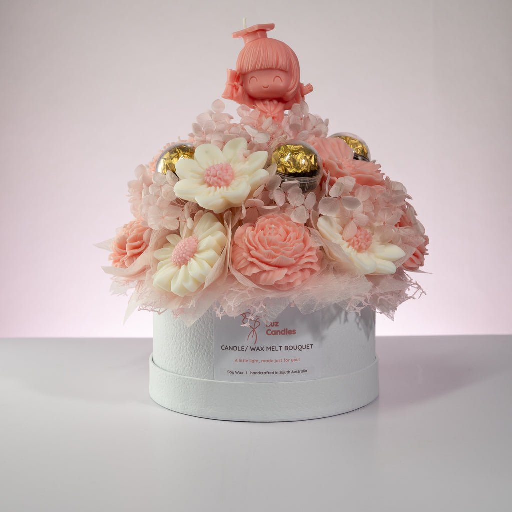 Luz Candles Au - A decorative candle and wax melt bouquet arranged in a round white gift box. The textured pink petals, gold foil-wrapped candies, and a small pink prom doll figure on top.