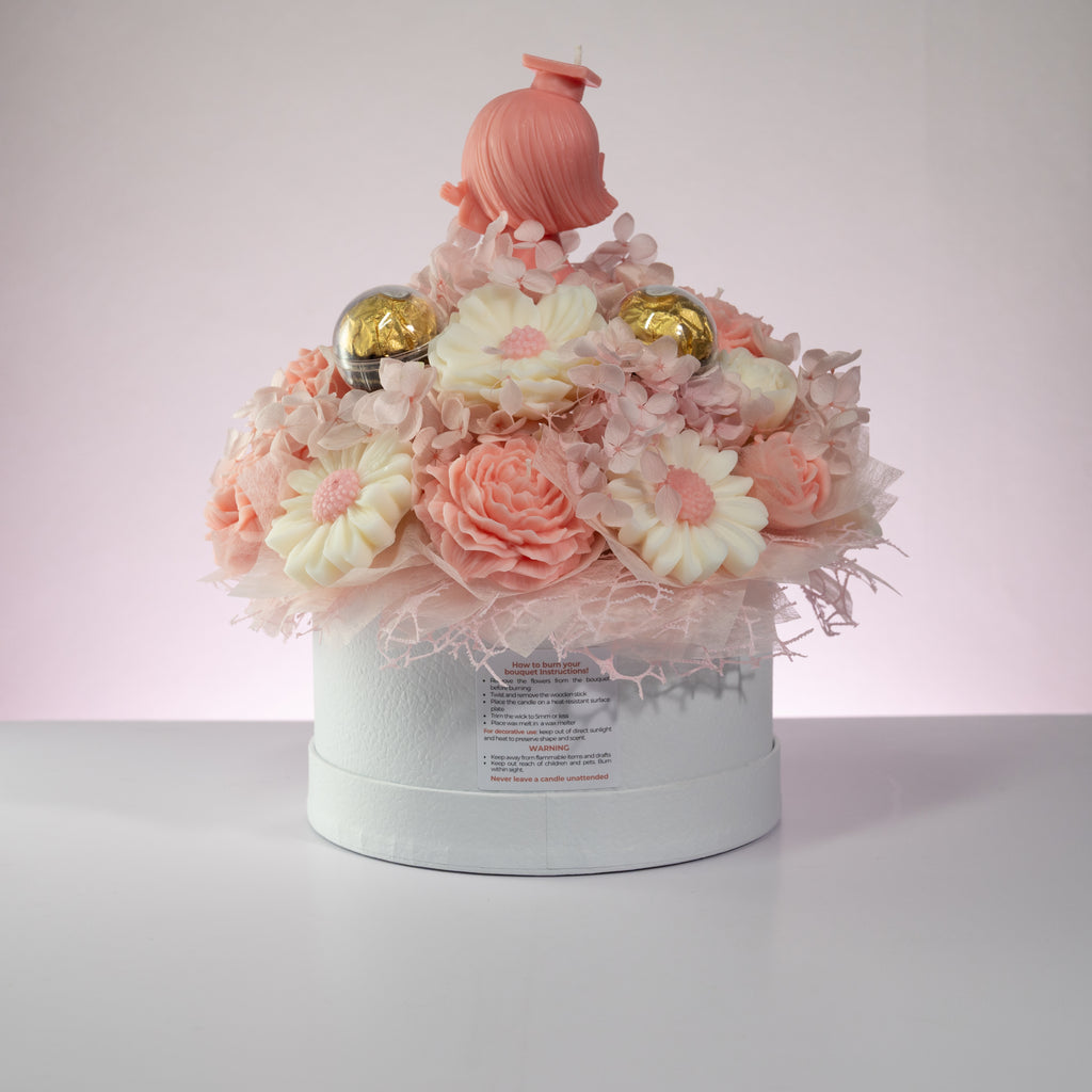 Luz Candles Au - A decorative round white gift box filled with pastel pink and white flower-shaped items, including daisy-like blooms, layered roses, small gold ornaments, and a pink hair-shaped topper, photographed on a light gray surface against a pale pink-lit background.