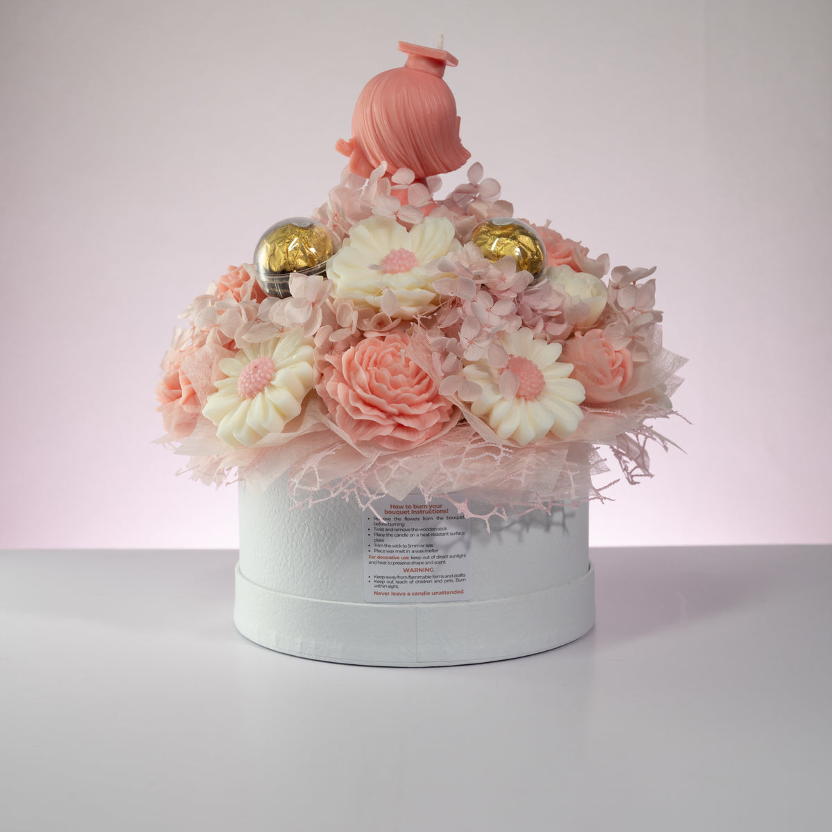 Luz Candles Au - A decorative round white gift box filled with pastel pink and white flower-shaped items, including daisy-like blooms, layered roses, small gold ornaments, and a pink hair-shaped topper, photographed on a light gray surface against a pale pink-lit background.