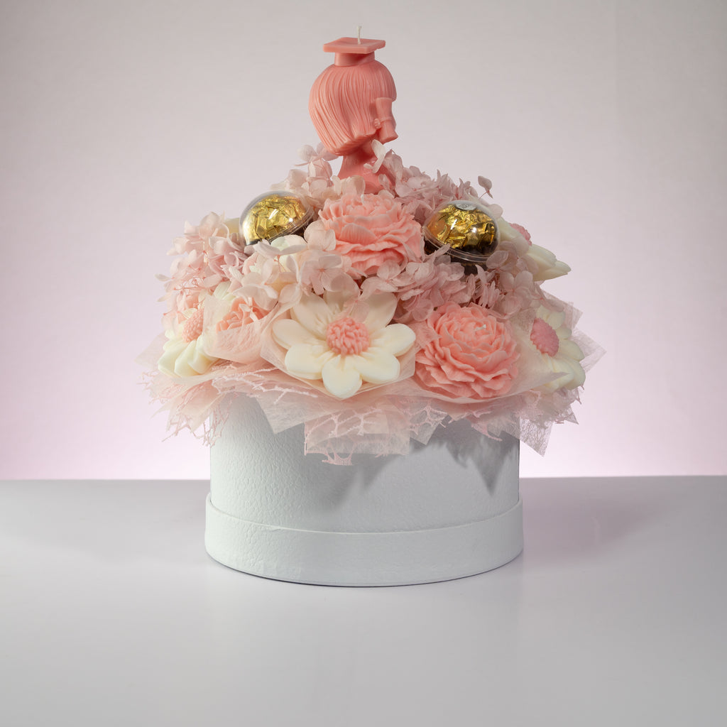 Luz Candles Au - A decorative floral arrangement in a round white box, featuring pink and white flowers, pink tulle, two gold foil-wrapped chocolates, and a pink hair-shaped topper on a light pink background.