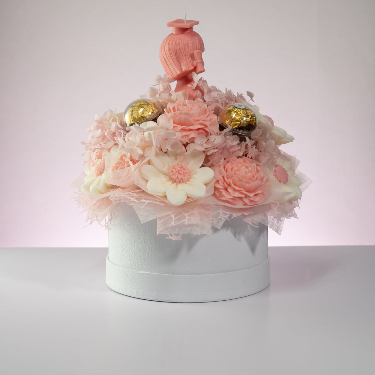 Luz Candles Au - A decorative floral arrangement in a round white box, featuring pink and white flowers, pink tulle, two gold foil-wrapped chocolates, and a pink hair-shaped topper on a light pink background.
