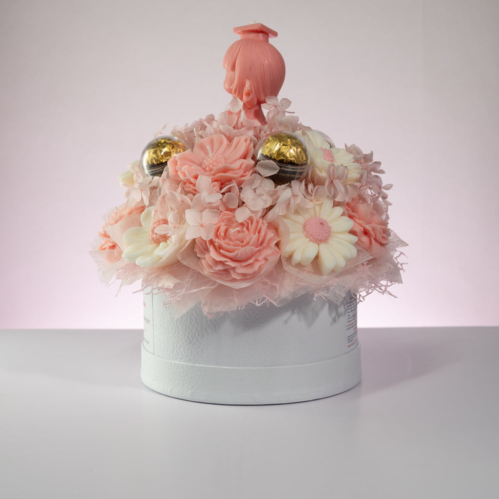 Luz Candles Au - A decorative round gift box filled with pastel pink and white flowers, two gold spherical ornaments, and a pink doll figure on top, photographed against a soft pink-lit neutral background.