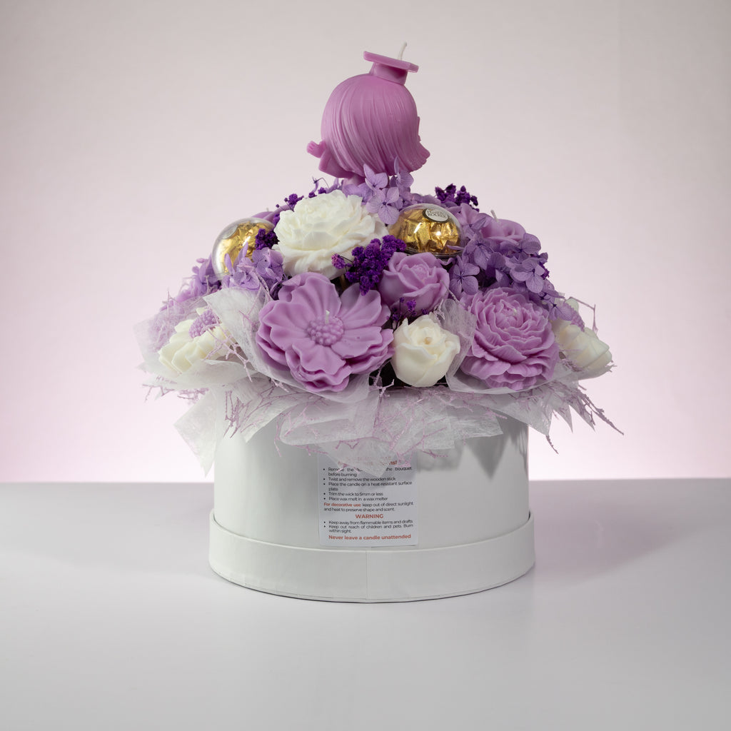Luz Candles Au - A round white gift box filled with a decorative floral arrangement in shades of lavender, purple, and white, topped with a pink hair doll candle, set against a soft pink and white studio background.
