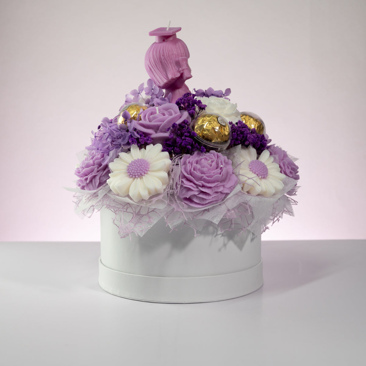 Luz Candles Au - A decorative round white box filled with purple and white artificial flowers, small purple berries, two gold chocolate eggs, and a pink figurine on top, photographed against a soft light pink studio background.