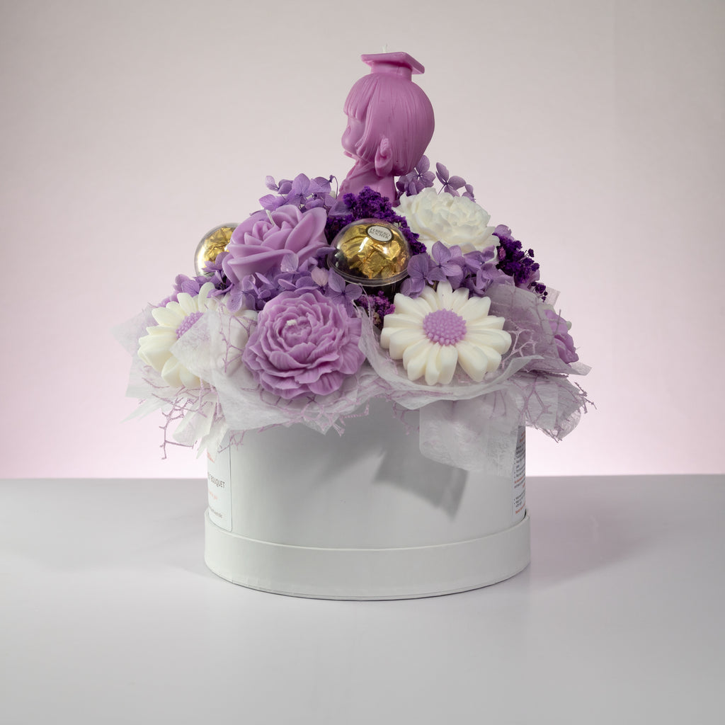 Luz Candles Au - A decorative gift box arrangement with purple and white flowers, two gold-wrapped chocolates, sheer white tulle, and a pink doll figure on top, presented in a round white box against a soft pink studio background.