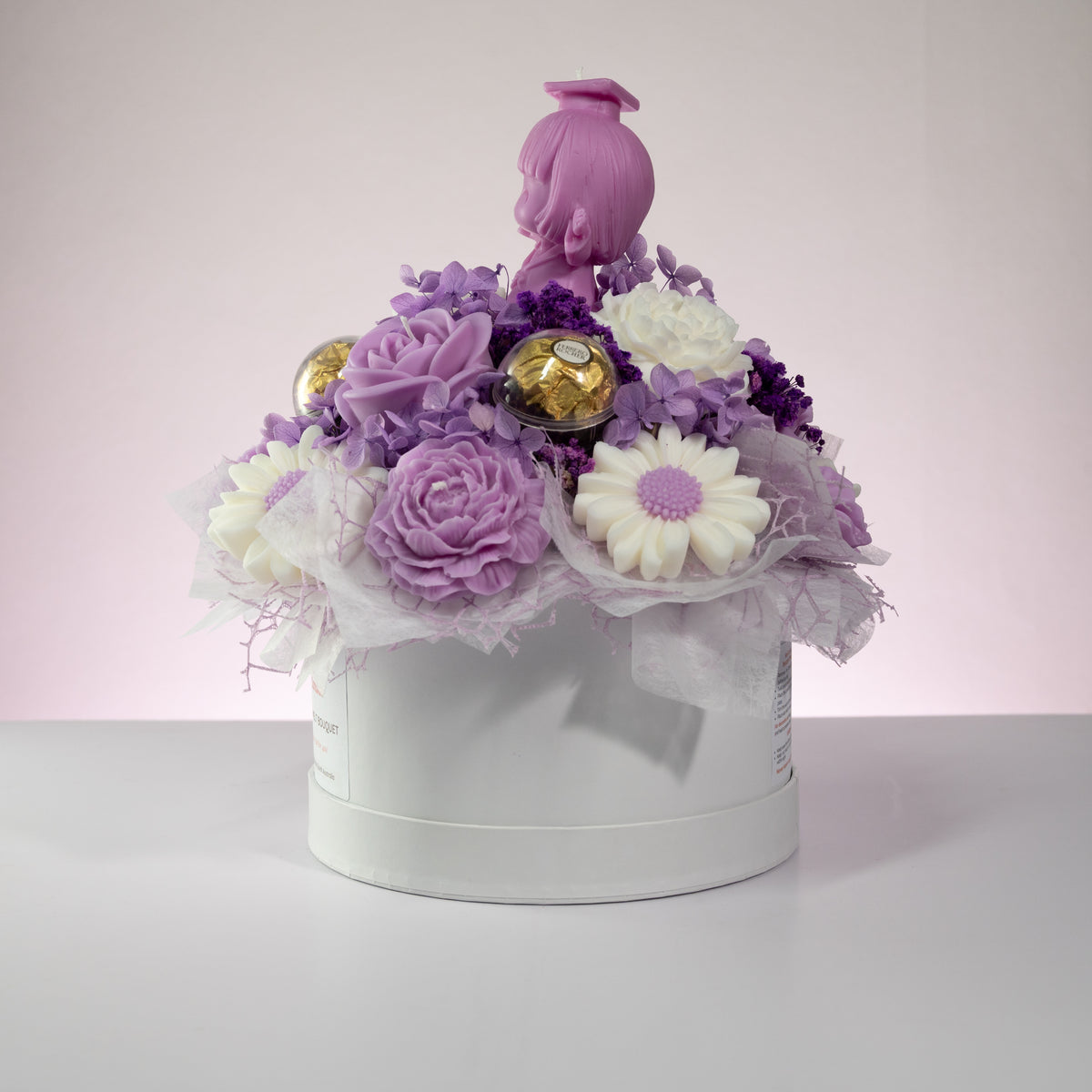 Luz Candles Au - A decorative gift box arrangement with purple and white flowers, two gold-wrapped chocolates, sheer white tulle, and a pink doll figure on top, presented in a round white box against a soft pink studio background.