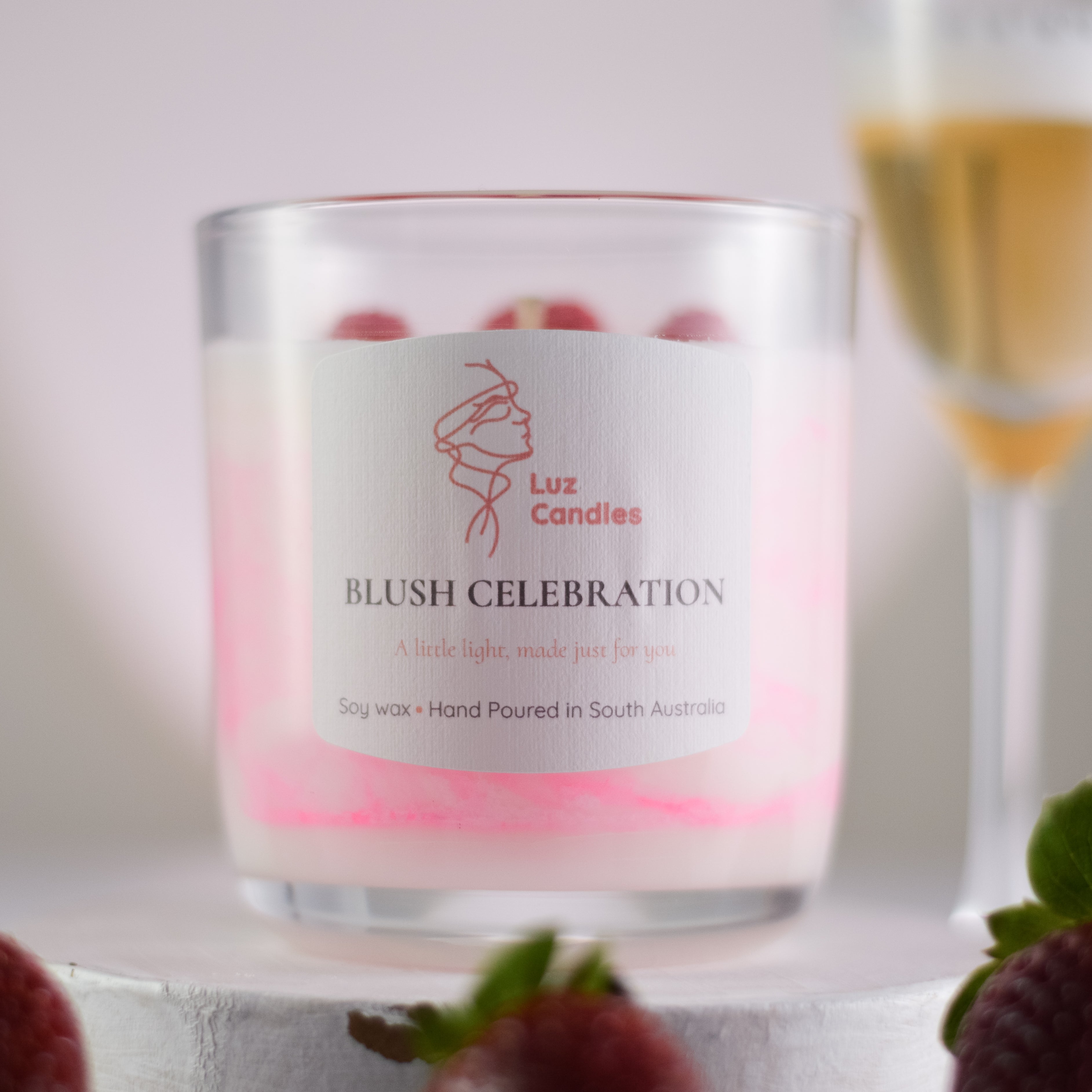 Luz Candles Blush Celebration candle with a blurred glass of wine and strawberries in the background.
