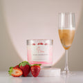 Candle labelled 'Blush Celebration' with strawberries and a glass of champagne on a neutral background