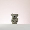 Luz Candles Au - A small grey koala figurine or candle centred on a plain light background, with a short white wick visible at the top and a textured koala body holding a wood stick. 