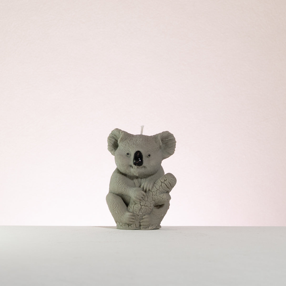 Luz Candles Au - A small grey koala figurine or candle centred on a plain light background, with a short white wick visible at the top and a textured koala body holding a wood stick. 
