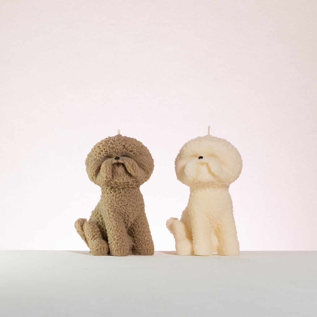 Two dog-shaped bichon frise candles, one brown and one white, on a light background