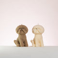 Two dog-shaped bichon frise candles, one brown and one white, on a light background