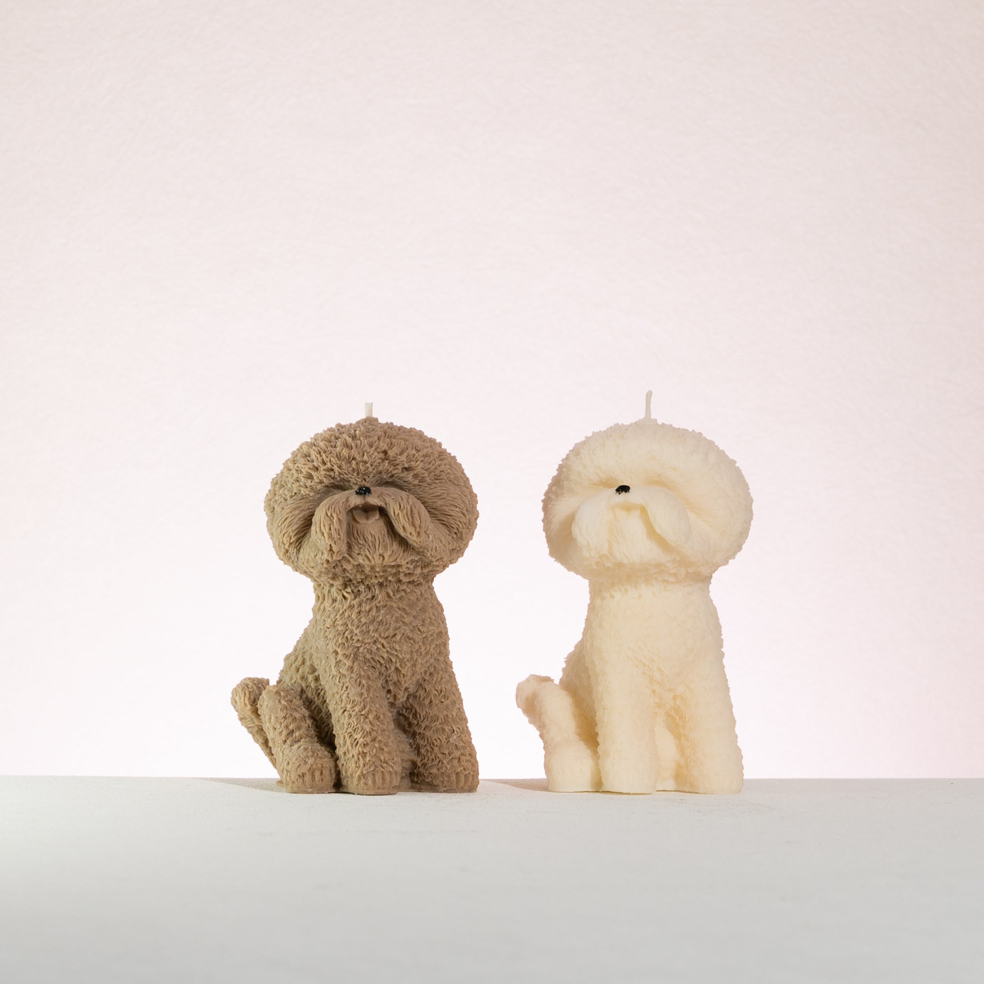 Two dog-shaped bichon frise candles, one brown and one white, on a light background