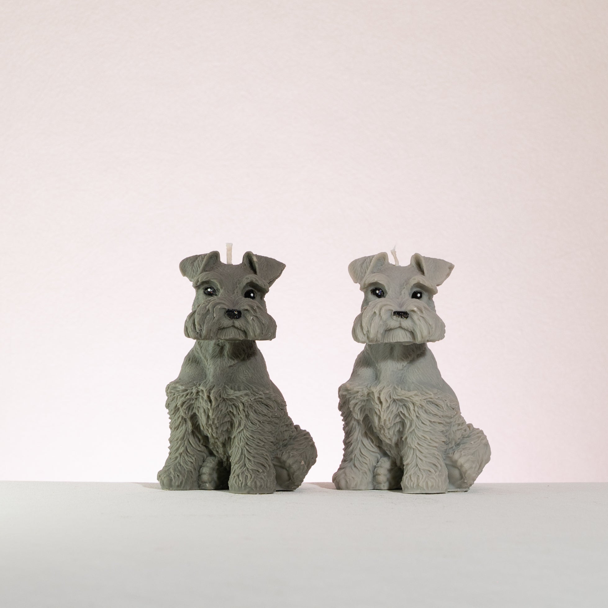 Two schnauzer dog-shaped candles on a white surface with a light pink background