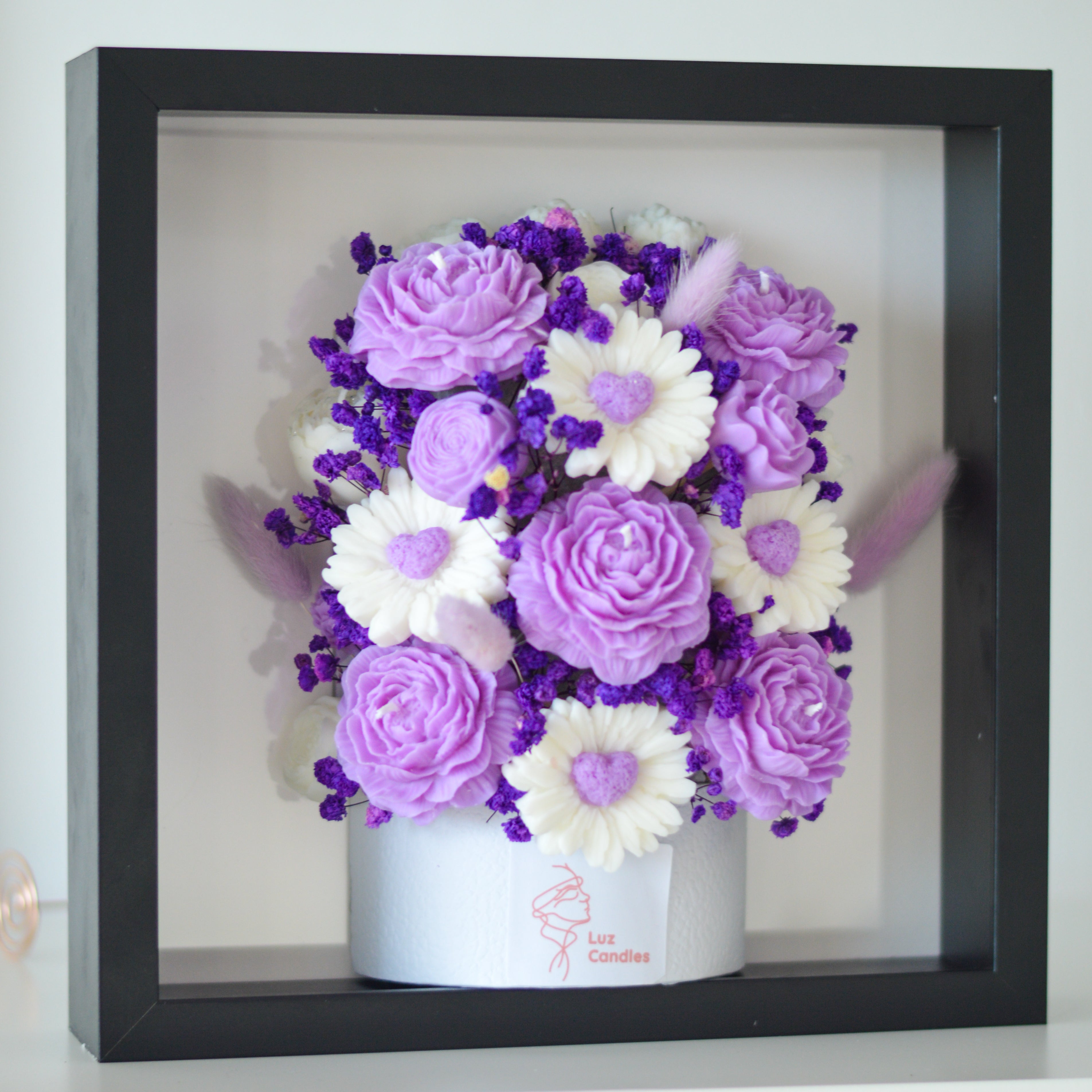 A framed floral arrangement featuring purple and white artificial flowers, displayed inside a black shadow box frame. The flowers include lavender-like roses, daisy-like blooms with purple centres, and small purple filler flowers, with a white base box printed with the brand name 'Luz Candles'.